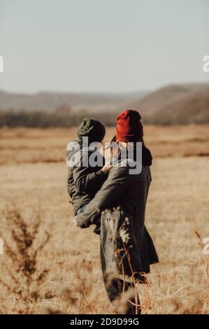 child in knitted warm green hat inhis mother arms in maroon hat , warmly hugging and admiring the autumn landscape of mountains together. Happy mother Stock Photo