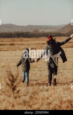 child in warm green knitted hat and his mother in maroon hat and warm coat, holding hands and running through the autumn field against the background Stock Photo