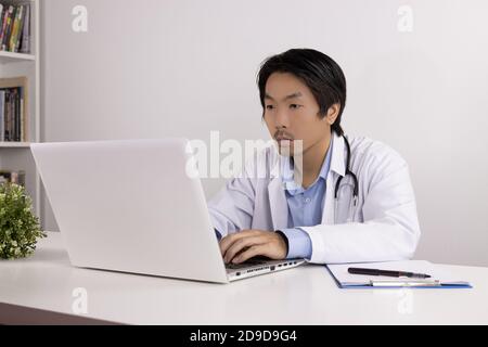 Young Asian Doctor Man in Lab Coat or Gown with Stethoscope Using Laptop Computer on Doctor Table in Office Stock Photo