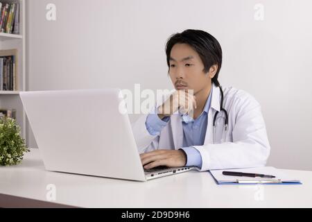 Serious Young Asian Doctor Man in Lab Coat or Gown with Stethoscope Touch Chin and Using Laptop Computer on Doctor Table in Office Stock Photo