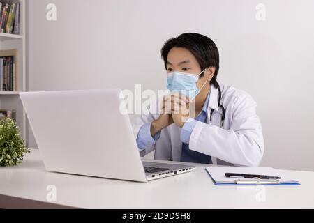 Young Asian Doctor Man in Lab Coat or Gown with Stethoscope Wear Face Mask and Thinking Front Laptop Computer on Doctor Table in Office Stock Photo