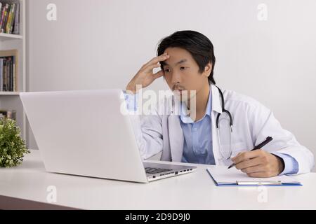 Serious Young Asian Doctor Man in Lab Coat or Gown with Stethoscope Writing Report and Using Laptop Computer on Doctor Table in Office Stock Photo