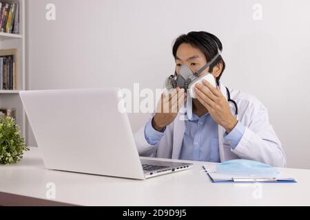 Young Asian Doctor Man in Lab Coat or Gown with Stethoscope Demonstrate Filter Mask Using with Patient Via Laptop Computer in Office Stock Photo