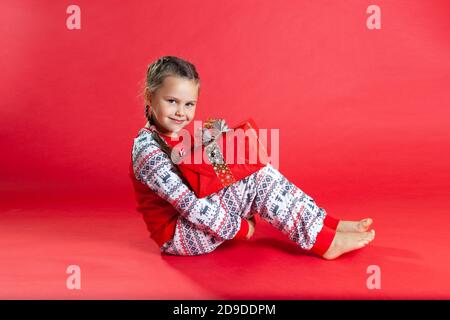 cute girl with two pigtails and in Christmas pajamas sitting on the floor, hugging her knees, holding a gift box Stock Photo