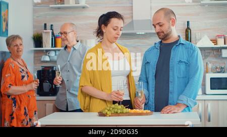 Handsome man making proposal to his girlfriend in front of his parents, in kitchen sitting near the table drinking a glass of white wine. Happy surprised woman kissing him while parents take a photo Stock Photo