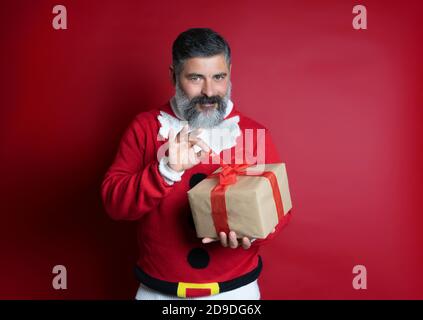 Portrait of a man who wears Christmas sweater opening gift box isolated on red background. Stock Photo