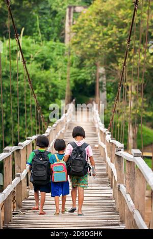 Asian primary school girls in casual clothes walking cross a simple suspension footbridge at dusk, rural scene near Thailand and Myanmar border. Stock Photo