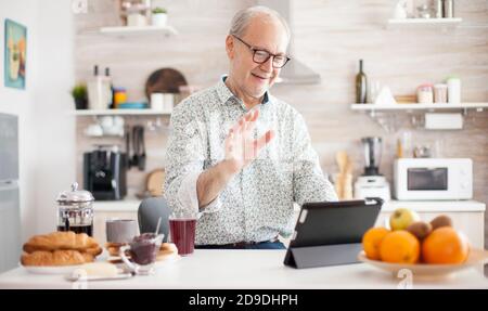 Authentic senior man on online video call in the morning in cozy kitchen. Elderly person using internet online chat technology video webcam making a video call connection camera communication conference call Stock Photo