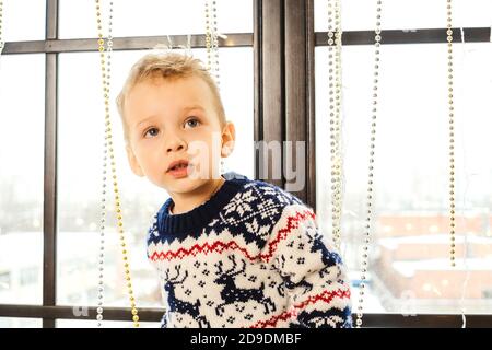 Sweet toddler boy in christmas sweater, standing near window and waiting for Satna Claus. Looking up. Stock Photo
