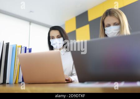Two women in protective medical masks sit at desk and work on laptops. Stock Photo