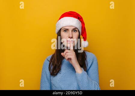 Portrait of cute young woman in Christmas red hat, makes hush gesture, dressed in winter blue sweater, poses against pastel yellow studio background. Stock Photo