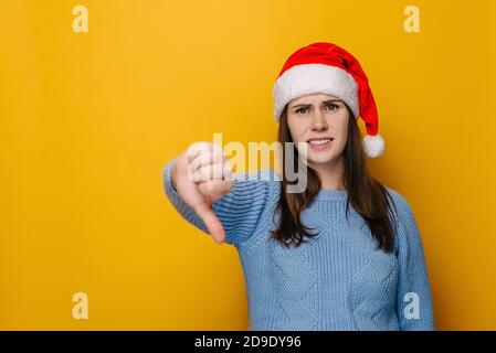 Discontent young woman shows disapproval sign, keeps thumb down, expresses dislike, frowns face in discontented, dressed in Christmas hat Stock Photo