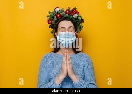 Young woman in traditional Christmas wreath hands folded in prayer beg about something good, wears respirator mask to protect herself from coronavirus Stock Photo