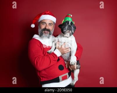 Portrait of man dressed for christmas holding a small french bulldog with elf hat on red background. Merry Christmas Stock Photo