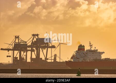 Cargo ship in front of a Dutch industrial plant with chimneys and windmill during sunset in Europoort, Rotterdam harbor Stock Photo