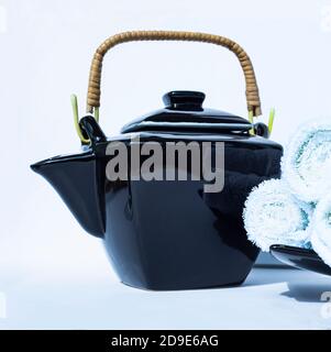 Black teapot with a wicker handle in oriental style and rolls of white terry towels on a white background. Stock Photo