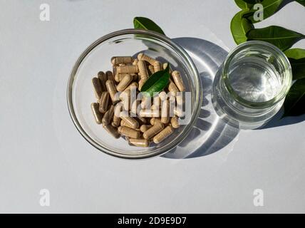 Glass bowl with capsules and glass jar with water on a white background. Green leaves. The idea of taking pills of supplements minerals vitamins. Stock Photo