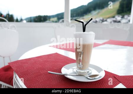 A glass cup of coffee latte with two plastic straws on a table outside. Serving caffeinated drinks in a cafeteria. Beautiful catering service. Stock Photo