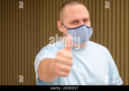 Upbeat positive young man wearing a reusable cloth face mask as protection during the Covid-19 pandemic giving a thumbs up gesture to show that all is Stock Photo