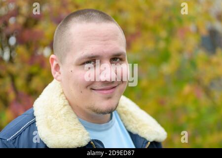 Relaxed young man wearing a faux fur trimmed casual jacket standing outdoors in autumn looking at the camera with a warm friendly smile Stock Photo