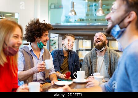 group of students having fun drinking coffee at coffeehouse wearing protective face mask.Young friends talking together at restaurant cafeteria. Stock Photo