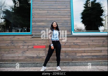 Hipster african american girl wearing jeans shirt with leopard sleeves posing at street against wooden house with windows. Stock Photo