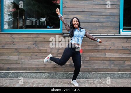 Hipster african american girl wearing jeans shirt with leopard sleeves jump at street against wooden house with windows. Stock Photo
