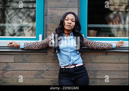 Hipster african american girl wearing jeans shirt with leopard sleeves posing at street against wooden house with windows. Stock Photo