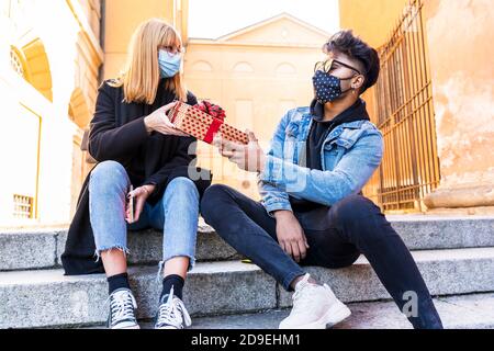 young multiethnic couple exchanges birthday presents wearing protective masks during the coronavirus emergency period Stock Photo