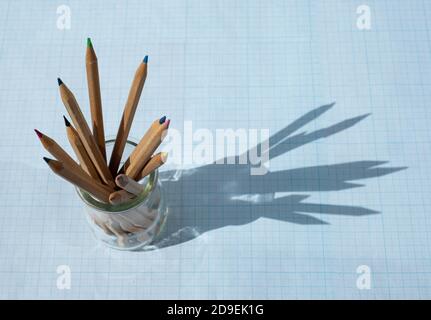 Colored wooden pencils in a glass beaker on a blue background. A shadow formed from the glass of pencils. Copy space for text. Stock Photo