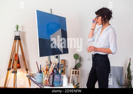A young painter with a glass of wine stands in her studio and observes her work of art on canvas. Besides you can see around her other accompanying ma Stock Photo