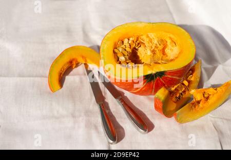 A ripe cut pumpkin and two steel kitchen knives on a woven napkin. Half pumpkin and wedges with seeds. Place for text on a light background. Stock Photo