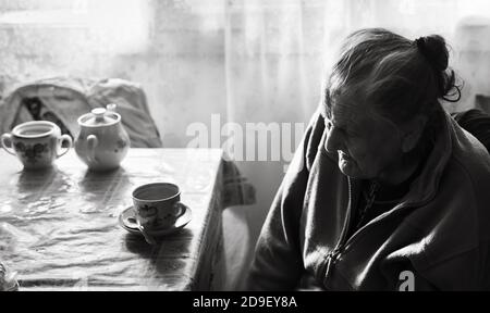 Black and white image of an old depressed woman. An elderly lonely woman sits at a table in the kitchen near the window and drinking tea. Stock Photo