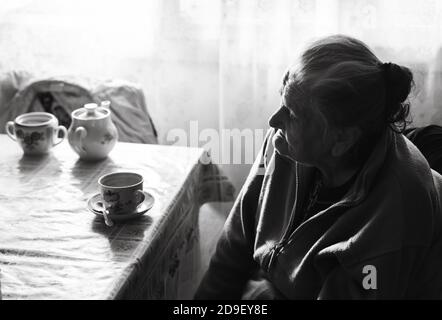 Black and white image of an old depressed woman. An elderly lonely woman sits at a table in the kitchen near the window and drinking tea. Stock Photo