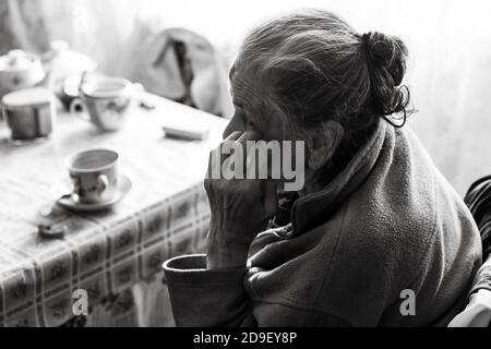 Old depressed woman. An elderly lonely woman sits at a table in the kitchen near the window and drinking tea. Stock Photo