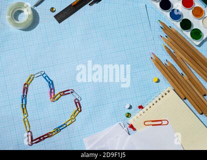 Stationery frame on blue graph paper. Paints pencils notepad paper and heart made of paper clips. Favourite buisness. Favourite hobby. Copy space  Stock Photo