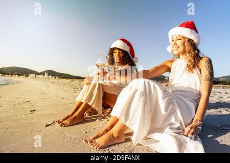 Tattooed caucasian young woman celebrating holidays with her black Hispanic girlfriend sitting on a seashore - Homosexual female couple toasting with Stock Photo