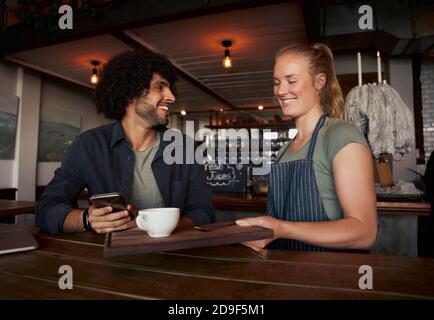 Cheerful female waitress serving coffee to young male customer with curly hair using smartphone in modern cafe Stock Photo