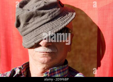 An elderly man in sunglasses relaxing on the sun while sitting on a red beach chair and covering his head with a cap. Lifestyle concept Stock Photo