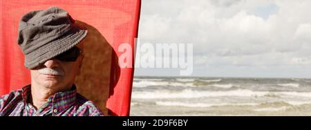 An elderly man sitting on a beach chair on the sea background covering his face by a cap. Lifestyle concept Stock Photo