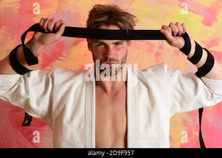 Man with confident face and bristle on colorful background. Karate fighter with fit strong body gets ready to fight. Guy poses in white kimono holding black belt. Healthy lifestyle and jujitsu concept Stock Photo