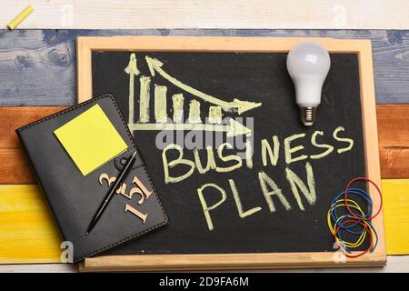 Business plan concept. Blackboard with business chart on colorful wooden background. Chalkboard with inscription written in yellow. Business decrease graph near lamp, organizer and other stationery. Stock Photo