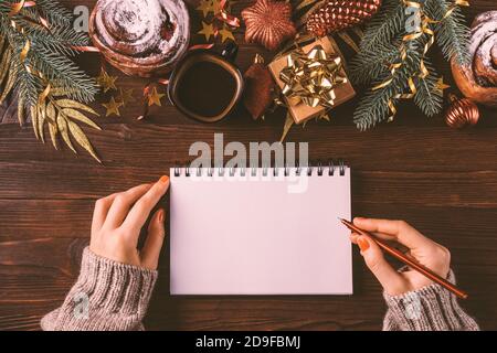Female hands writing in blank notebook among Christmas decorations, fir branches and cup of coffee with bun on dark table, top view. Stock Photo
