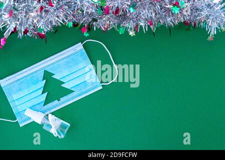 Medical face shield with cut out silhouette of Christmas tree and hand sanitizer on green background. With tinsel near the mask. Christmas and New Year concept, coronavirus concept. Stock Photo