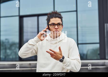 Handsome young man with curly black hair is on the street against building talking by the phone Stock Photo
