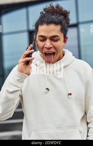 Handsome young man with curly black hair is on the street against building talking by the phone Stock Photo