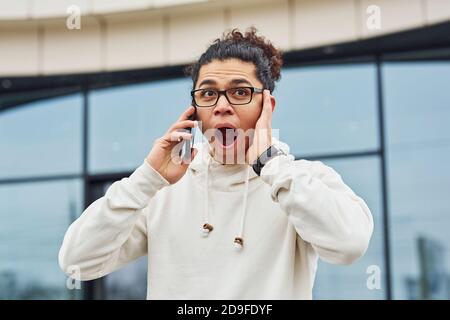 Handsome young man with curly black hair is on the street against building talking by the phone Stock Photo