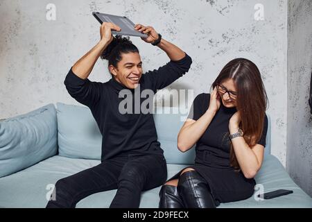 Man with curly hair and woman that in black clothes sitting with laptop indoors. Guy is angry Stock Photo