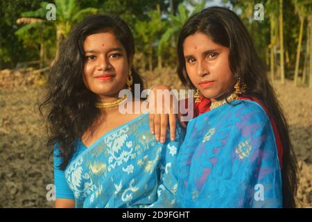 Two beautiful Indian Bengali Teenage girls wearing blue sari, necklace earrings nose pin posing in a plowed agricultural field one hand on shoulder. Stock Photo