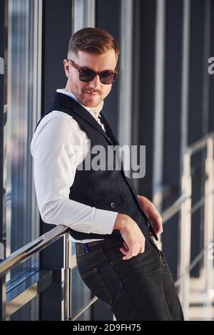 Man in elegant clothes and sunglasses is standing indoors and posing for the camera Stock Photo